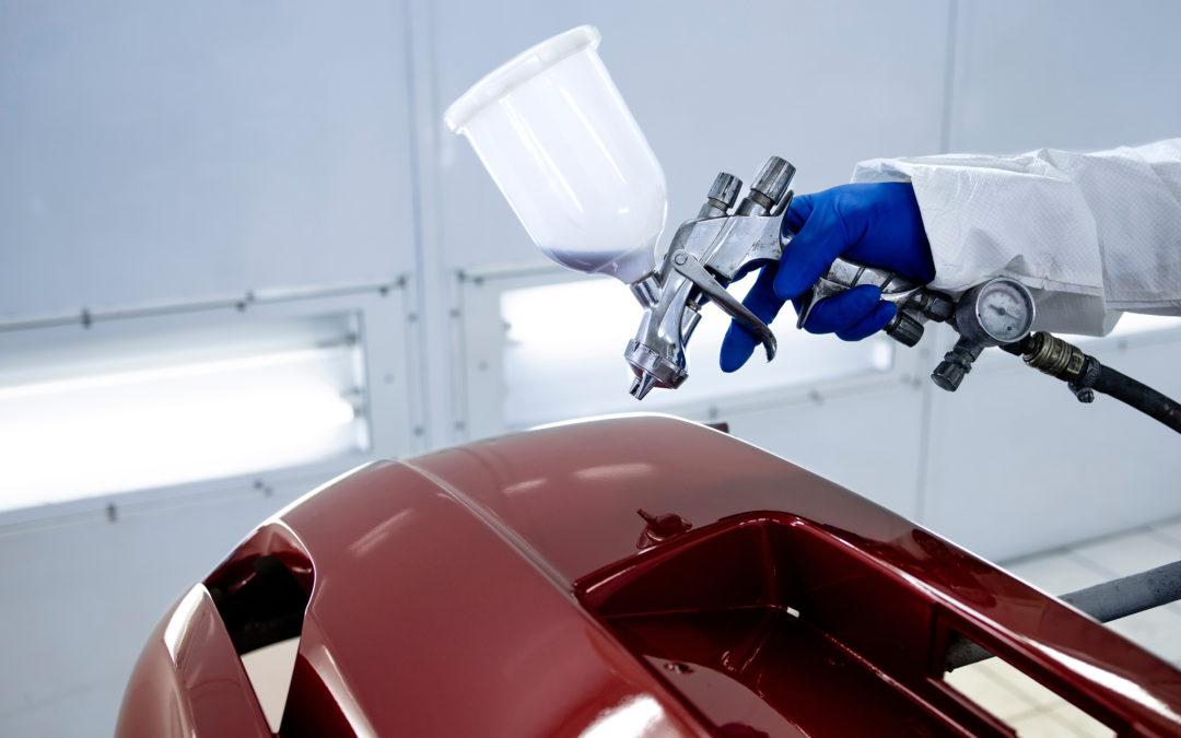 Car painter in protective clothes and mask painting automobile bumper with metallic paint and varnish in chamber workshop.